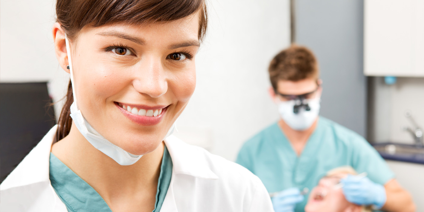 Female Dental Assistant smiling. A male Dental Assistant is conducting a cleaning behind her