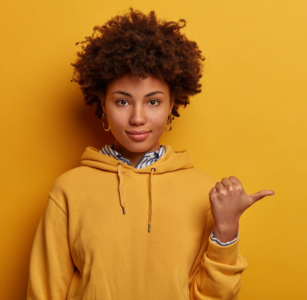 Calm lovely Afro American woman points thumb aside, shows nice place to visit, wears hoodie, isolated on yellow studio background, suggests good offer or proposal