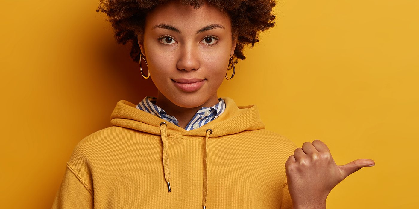 Calm lovely Afro American woman points thumb aside, shows nice place to visit, wears hoodie, isolated on yellow studio background, suggests good offer or proposal