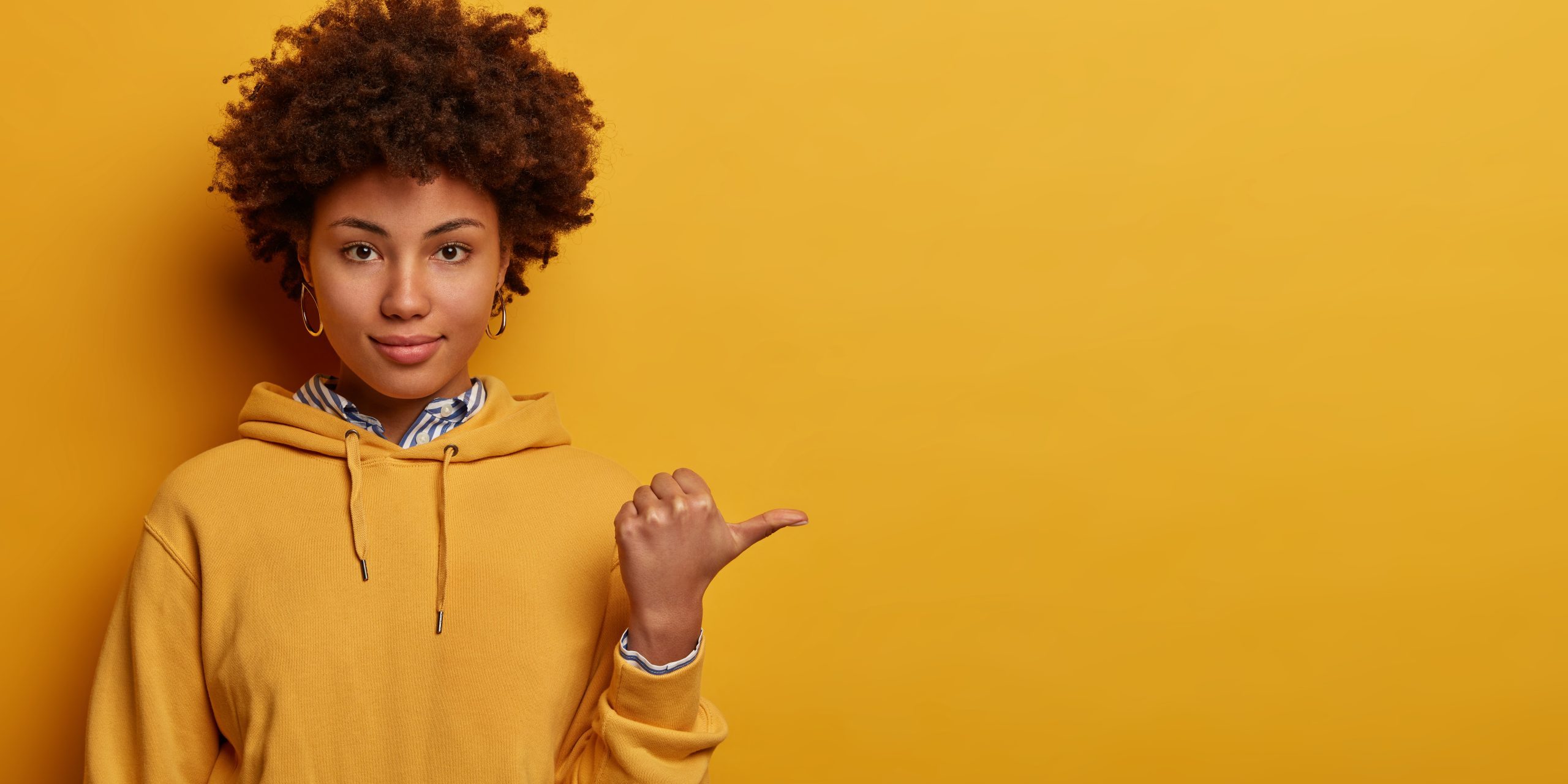 Calm lovely Afro American woman points thumb aside, shows nice place to visit, wears hoodie, isolated on yellow studio background, suggests good offer or proposal.
