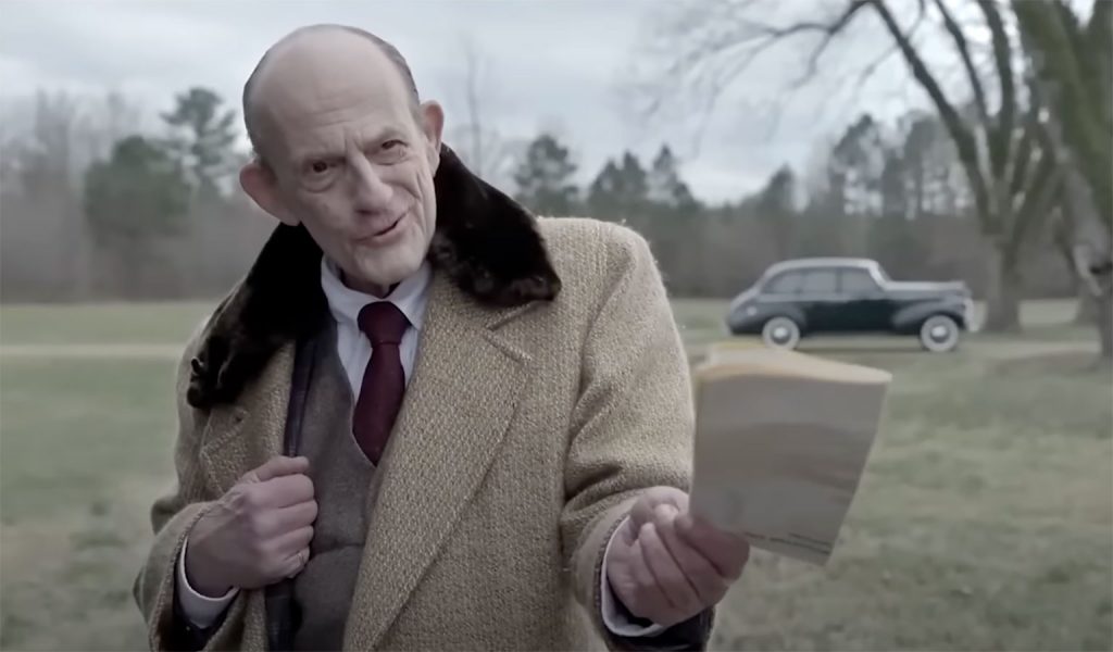 Christopher Lloyd in suit holding paper toward someone off-screen.