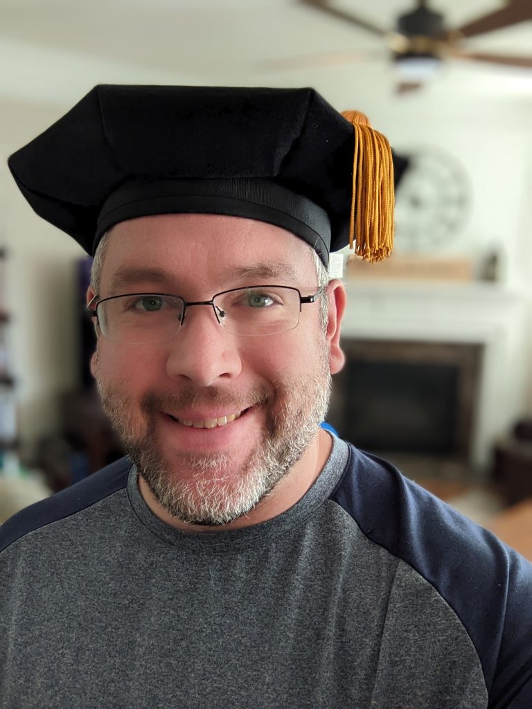 white man with graying beard wearing PhD tam and baseball tshirt