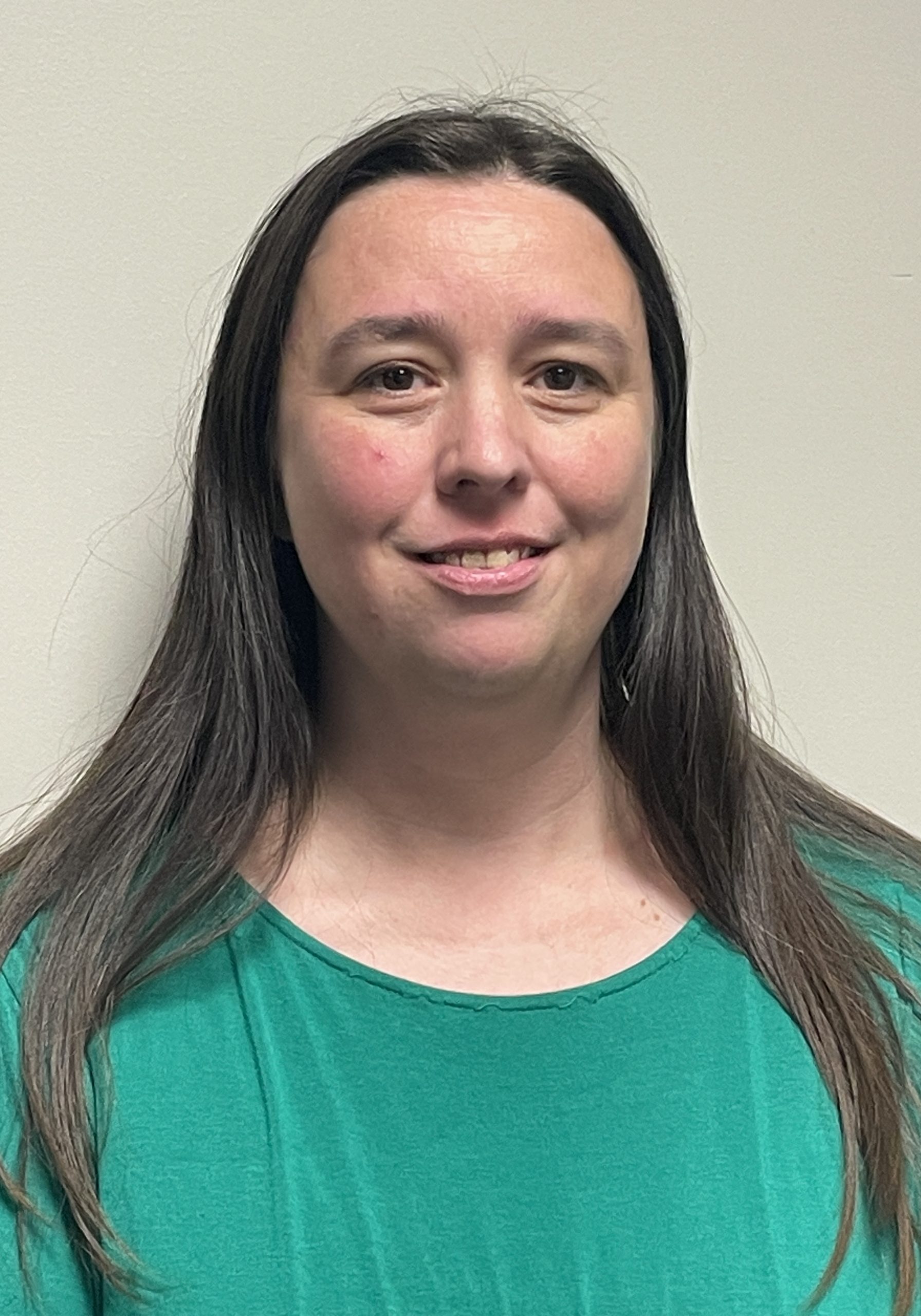 Smiling woman with long, straight Brown hair and wearing a green top in front of a plain white background.