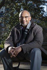 Photograph of an African-American man seated outside on a park bench wearing a winter coat and scarf. Photo credit is to Lauren Kress