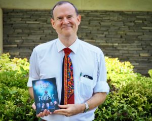 Man in sleeves and tie displaying a novel cover and smiling