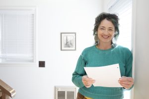 Woman in a turquoise sweater leaning against a wall, holding some papers, smiling with white space to the left of her